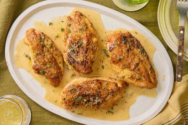 Lemon Pepper chicken on a white platter on a mustard colored textile, with yellow napkin, plates and drinks surrounding it