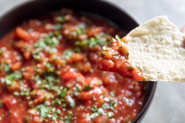 Close-up of a chip topped with salsa. A bowl is in the background, slightly out of focus.