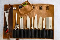 An open Rustic Town Genuine Leather Chef Knife Roll containing assorted kitchen knives a sharpening rod a grater a slotted spoon and a few markers
