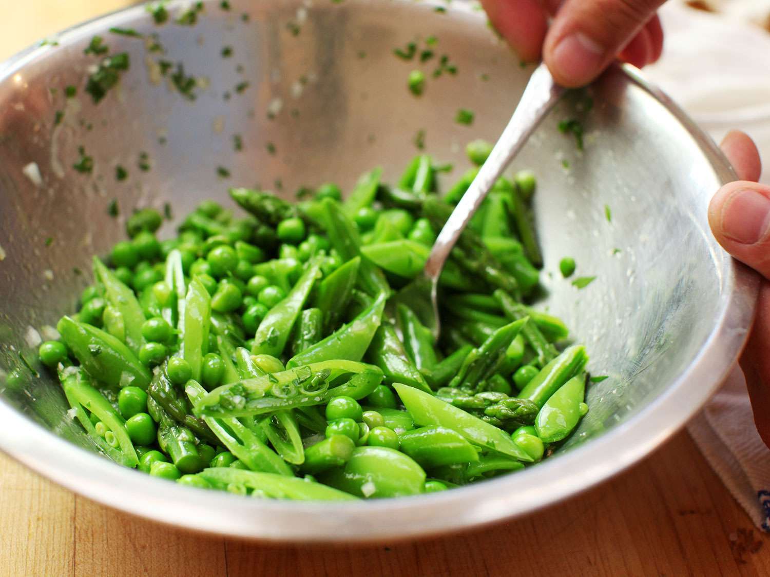 The peas, favas, and other blanched vegetables are tossed with dressing in a mixing bowl.