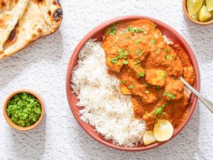A bowl of tofu tikka masala with rice and garnished with fresh herbs accompanied by naan bread lime wedges and a small bowl of chopped herbs