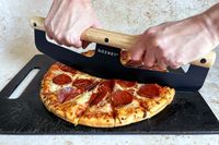 Hands slicing a pepperoni pizza using a Gozney pizza rocker on a cutting board