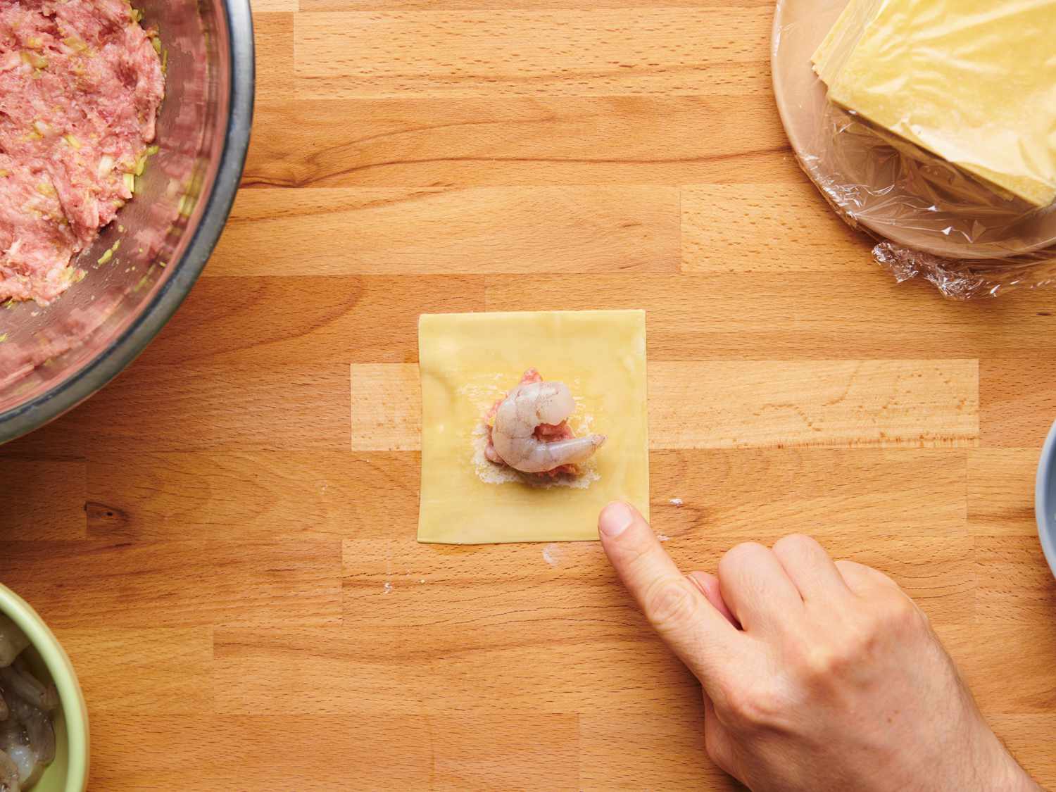 A fingertip moistening a wonton wrapper with water around the edge. Pork filling is in the center of the wonton and is topped with a single shrimp.