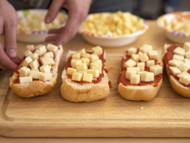 Several split french bread on a cutting board, slathered with tomato sauce and topped with different brands of cubed low-moisture mozzarella.