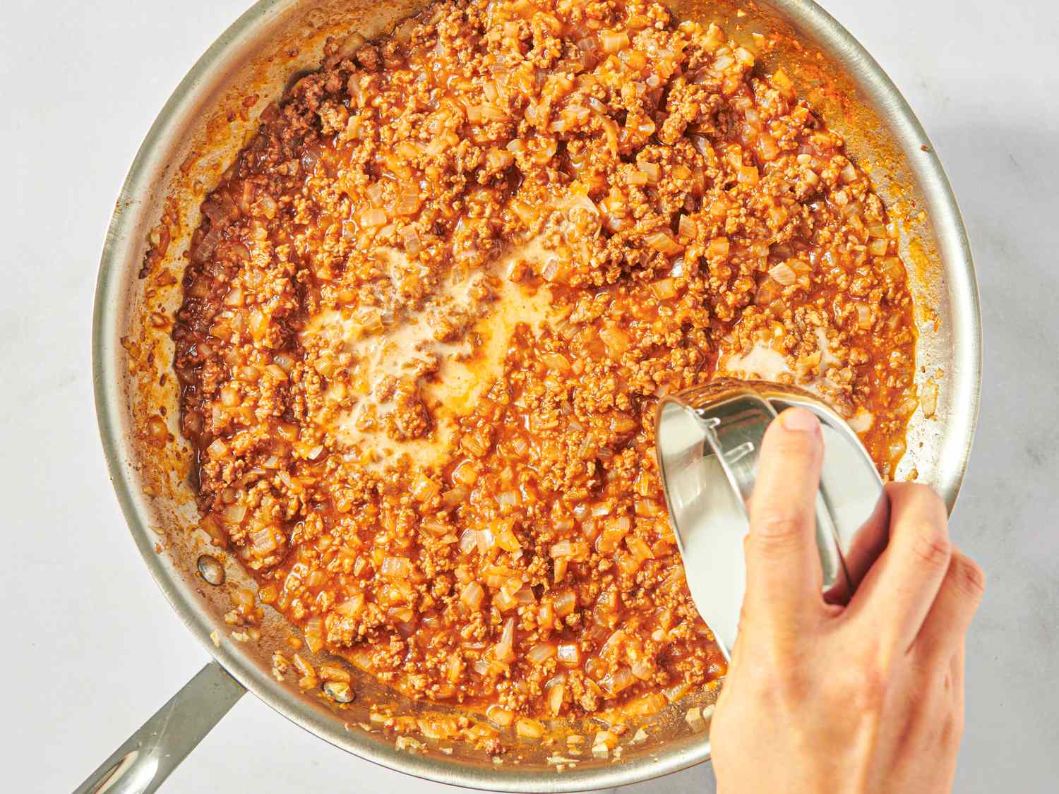 A skillet with a sloppy joe mixture being stirred with a spoon by hand