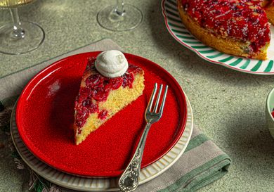 A slice of cranberry upsidedown cake on a red plate with a dollop of cream fork placed alongside the slice whole cake in background