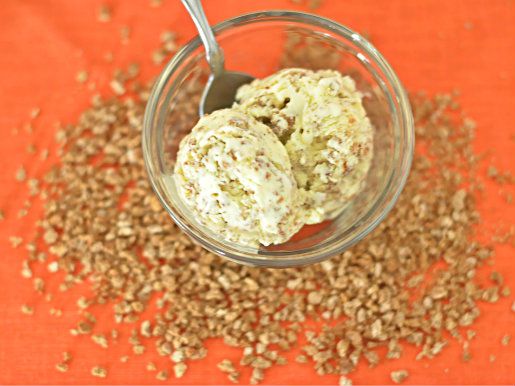 Overhead view of two scoops of Grape-Nuts ice cream served in a small bowl. Grape-Nuts are scattered on the orange surface below.