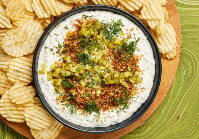 Bowl of pickle dip garnished with panko, dill and pickles, surrounded by a wooden plate full of potato chips, on a green tabeltop