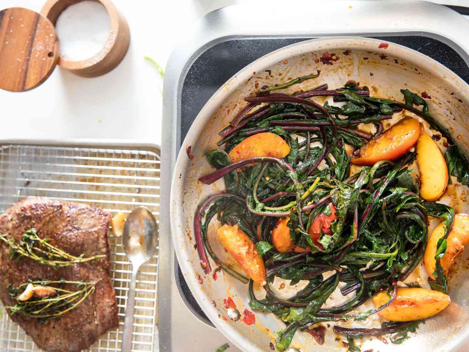 Overhead shot of resting flank steak on a wire rack, next to a skillet full of wilted greens and seared peach wedges on an induction burner.