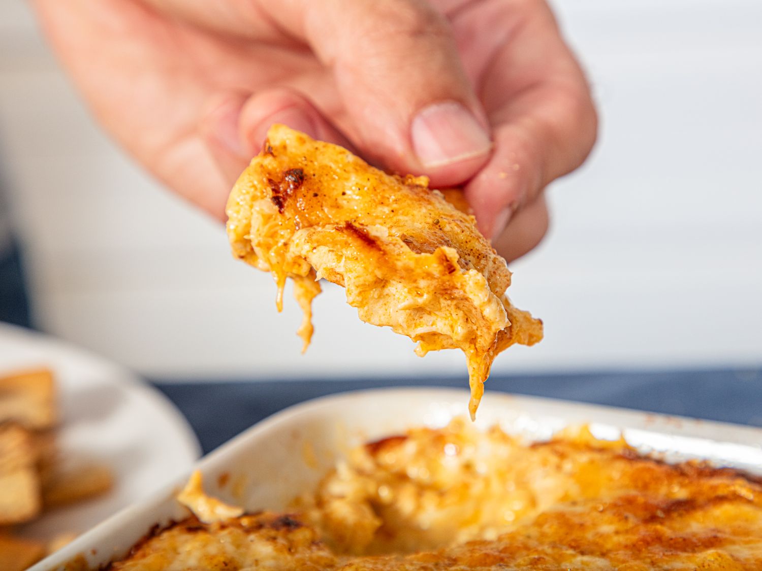 A hand holding a cracker with cheese and dip scooped from a dish of baked dip