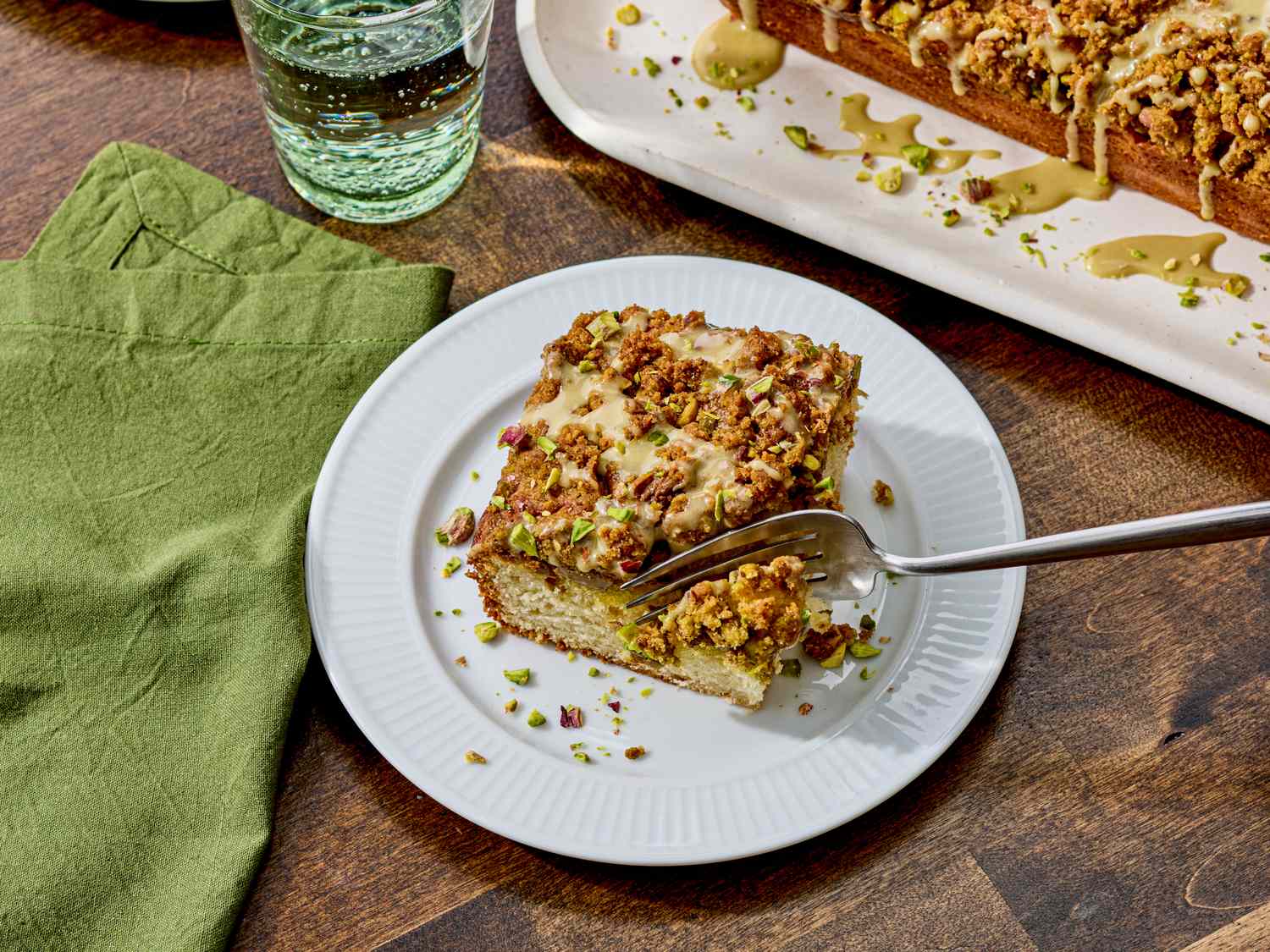 A slice of pistachio coffee cake on a plate with a fork a green napkin and glass of water nearby tray with more cake in the background