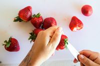 A person holding a strawberry while cutting its stem with a Material knife