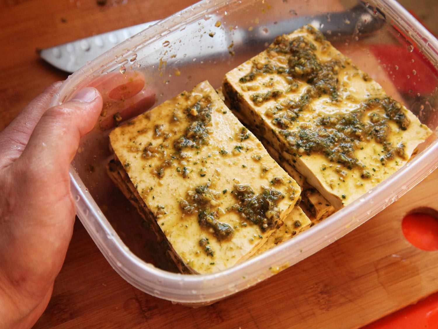 Two piles of sliced tofu in a container with marinade. A hand is holding the container and a knife is in the background.
