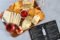 Cheese, crackers and a small bowl of peppers from Curdbox Cheese Subscription Box on a wooden board alongside a jar of red jam and a pairing menu sheet