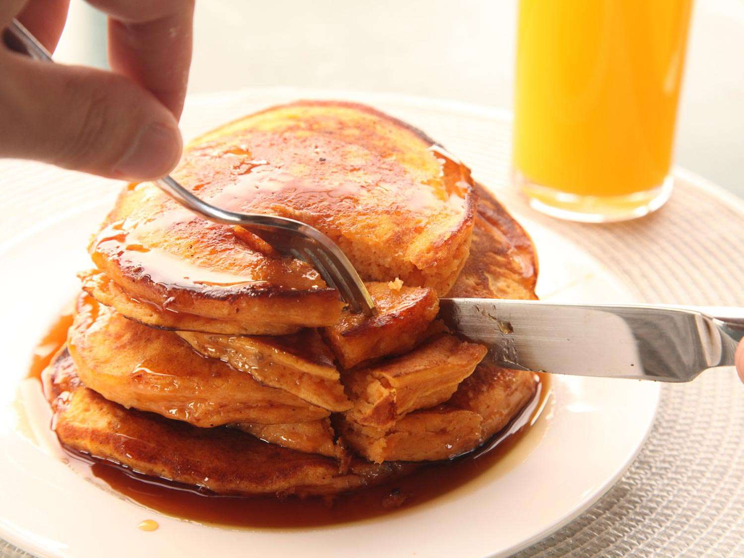 Hands with a fork and knife cutting into a stack of sweet potato pancakes with maple syrup.