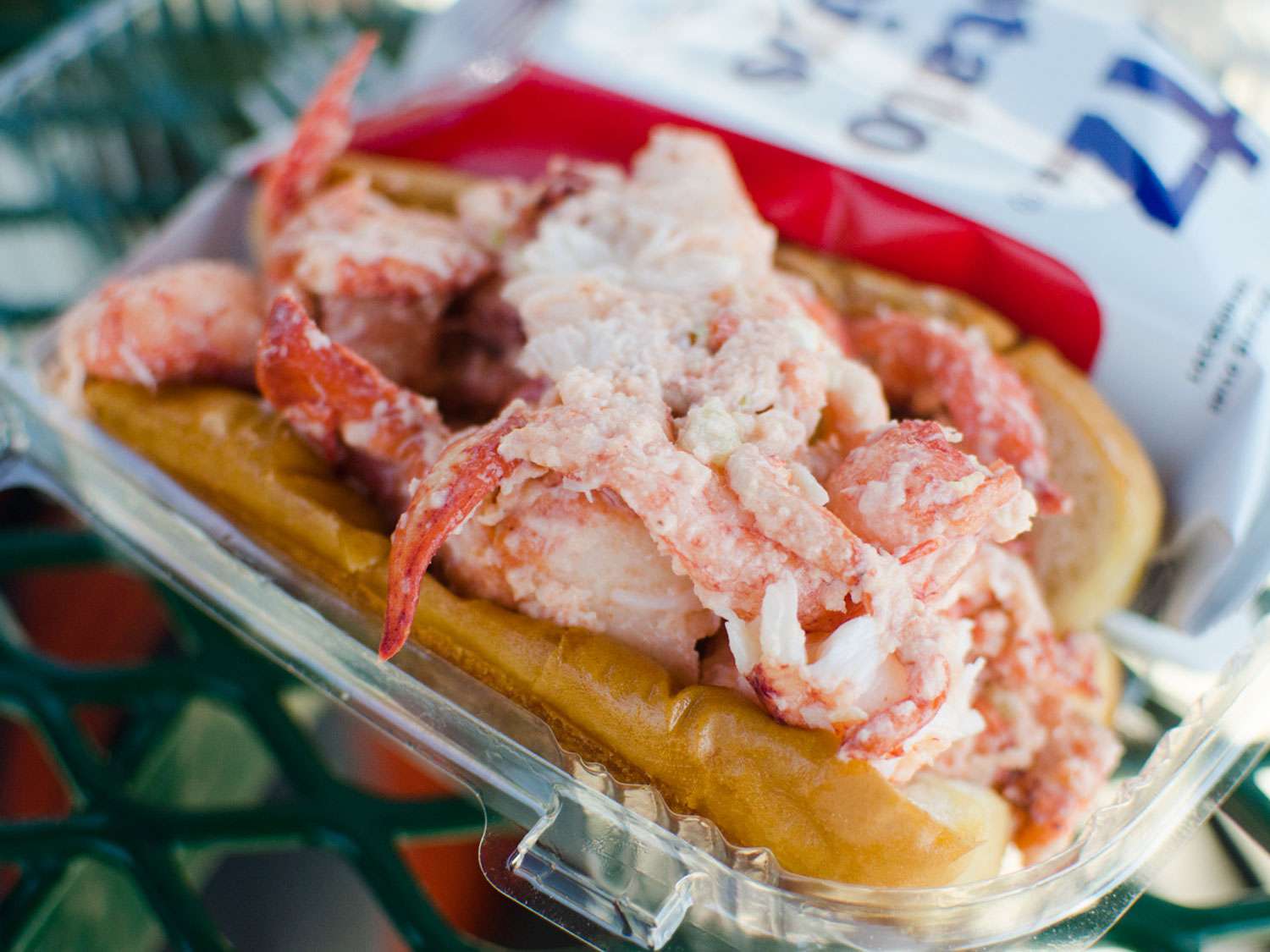 A lobster roll in a plastic container with a bag of chips.