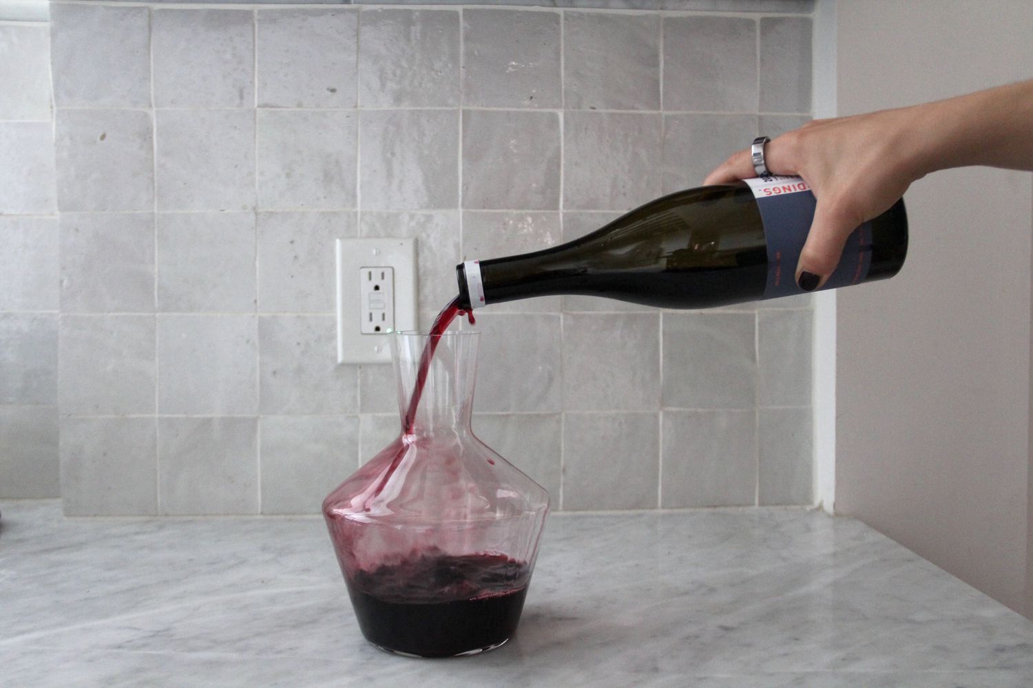A person pours a bottle of red wine into a wine decanter on a marble counter