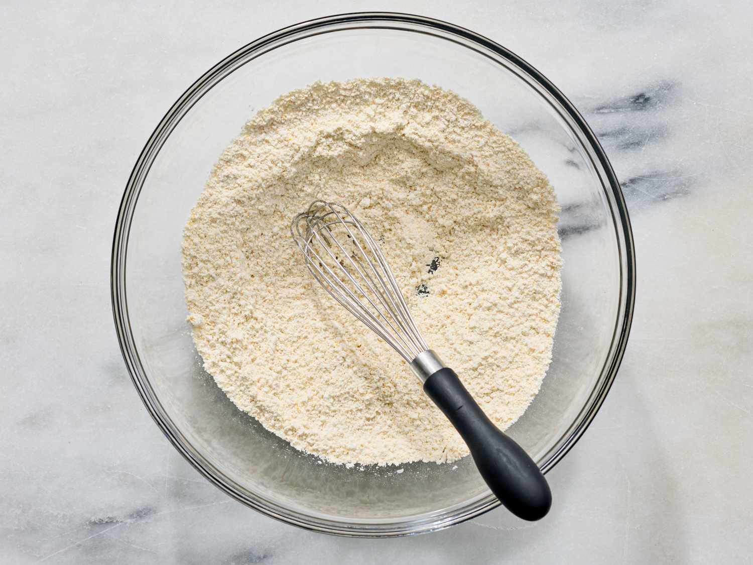 A glass bowl on a marble surface containing dry ingredients with a whisk placed inside
