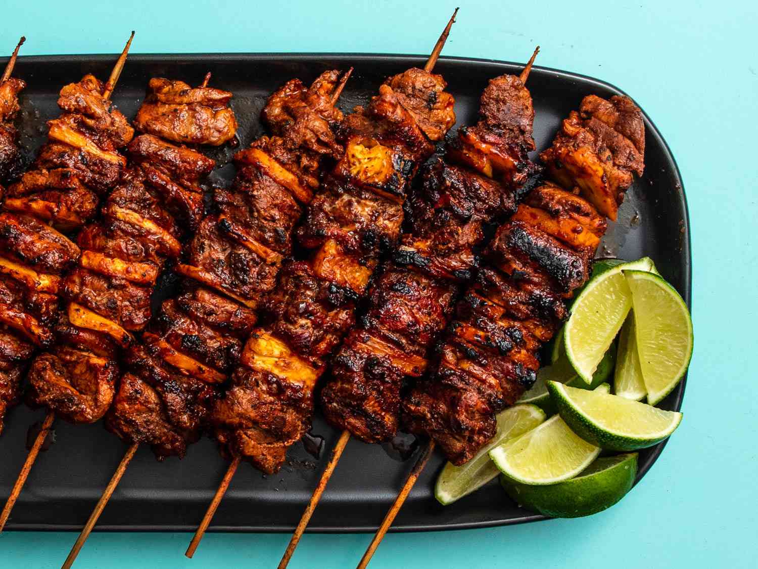 Grilled al pastor pork skewers on black platter with lime wedges