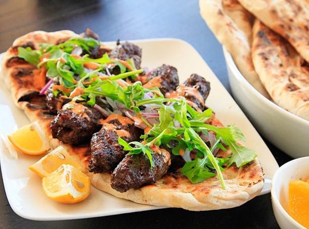 Kofte (minced-lamb) kebabs laid out on flatbread, topped with spicy harissa-yogurt sauce and arugula