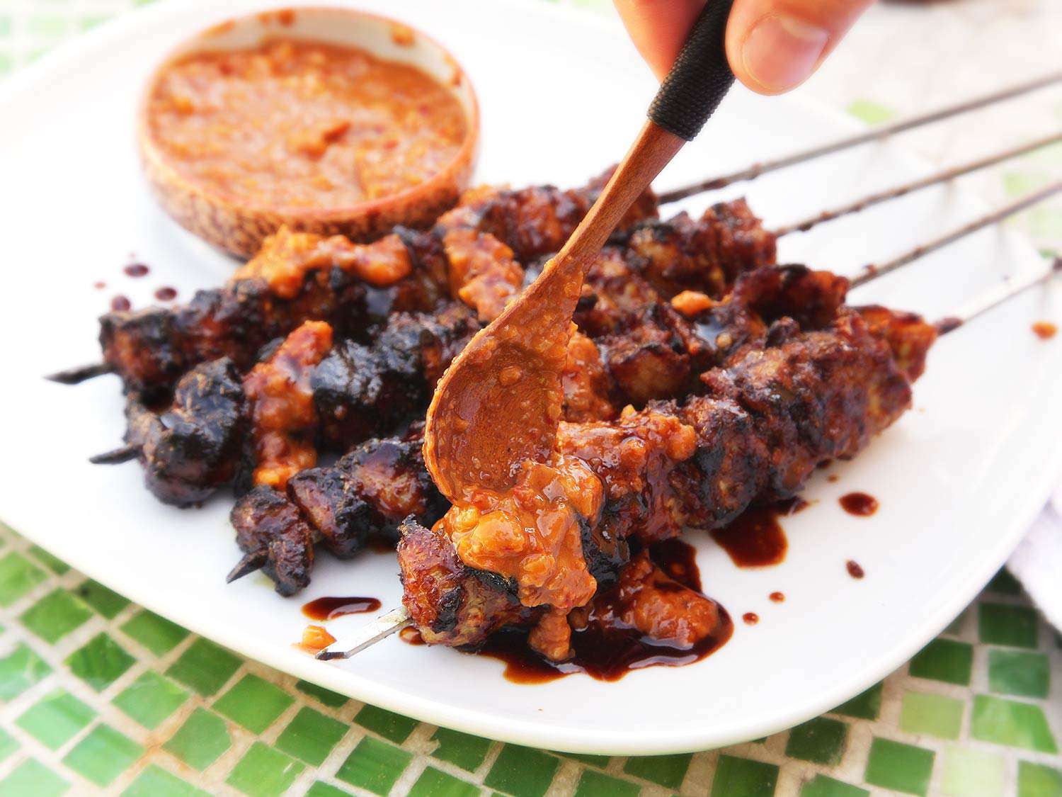 Spooning peanut sauce over skewered pork satay on a white plate.