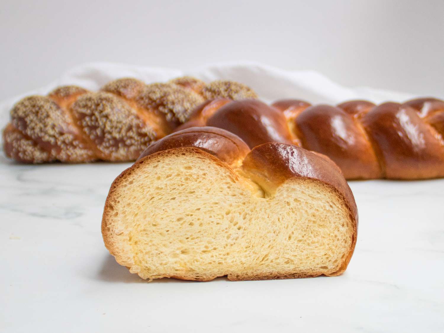 a slice of challah standing up on a marble surface. 2 loaves of challah are in the backgorund, one is plain, one has sesame seeds 