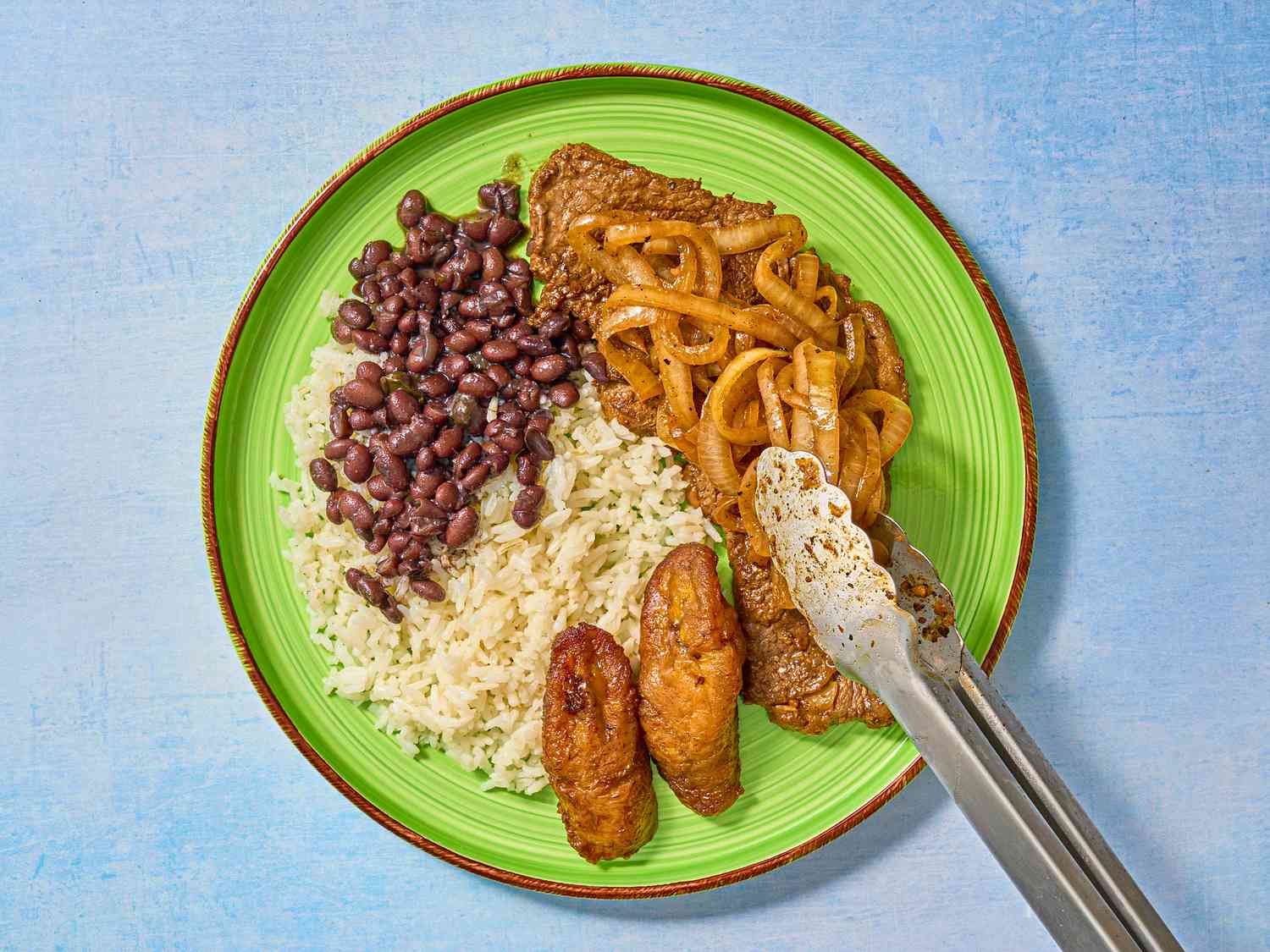 Onions placed on top of steaks, with sides of rice, plantains and beans 
