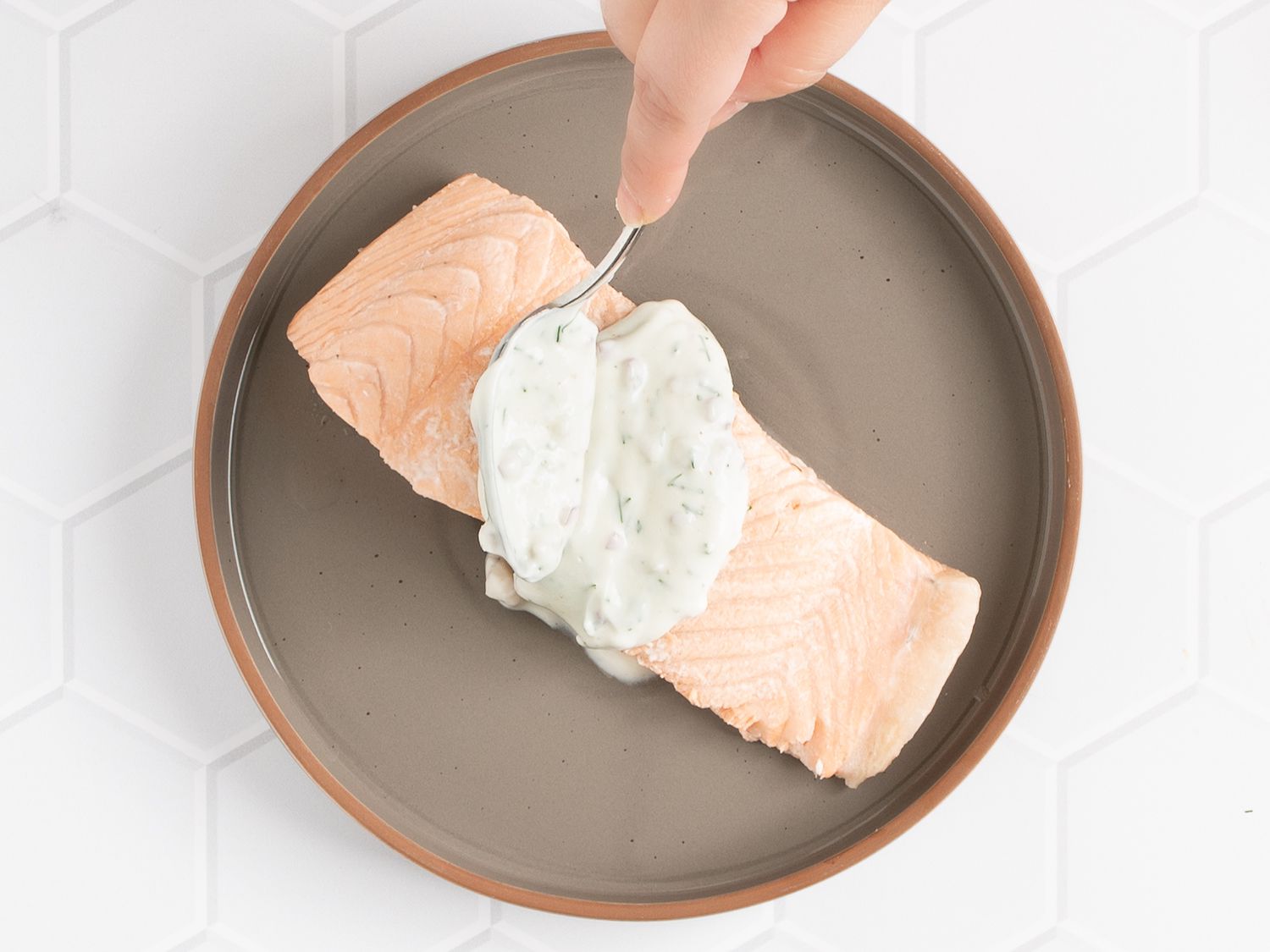 A hand spooning yogurt sauce on top of a poached salmon filet.