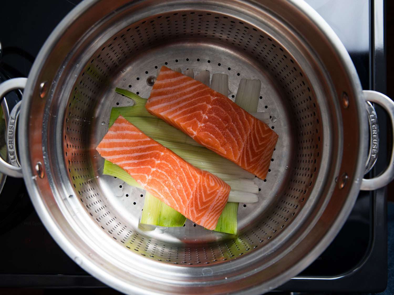 Overhead shot two pieces of salmon fillet nestled in a steamer insert on top of a bed of leek greens.