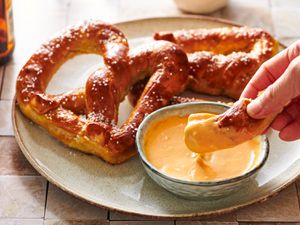 A round ceramic plate holding two Bavarian-style pretzels and a small bowl of beer cheese. Part of one pretzel is being dipped into the beer cheese by a hand.