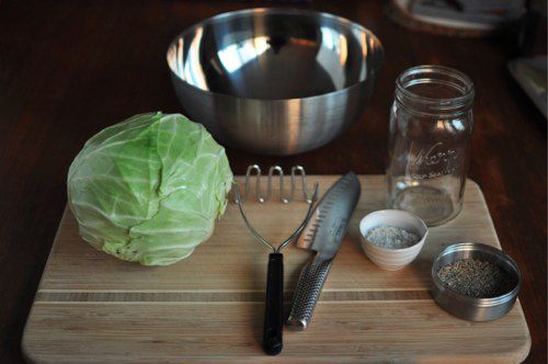 Everything needed to make sauerkraut, displayed on a cutting board: mixing bowl, quart-sized canning jar, potato masher, kitchen knife, cabbage, salt, caraway seed.