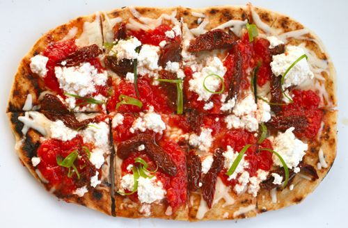 A grilled pizza topped with cheese, tomato sauce, ricotta, sun-dried tomatoes, and shreds of basil.