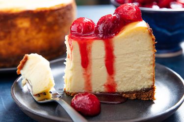 A slice of epic New York Cheesecake on a plate, topped with cherry sauce. 