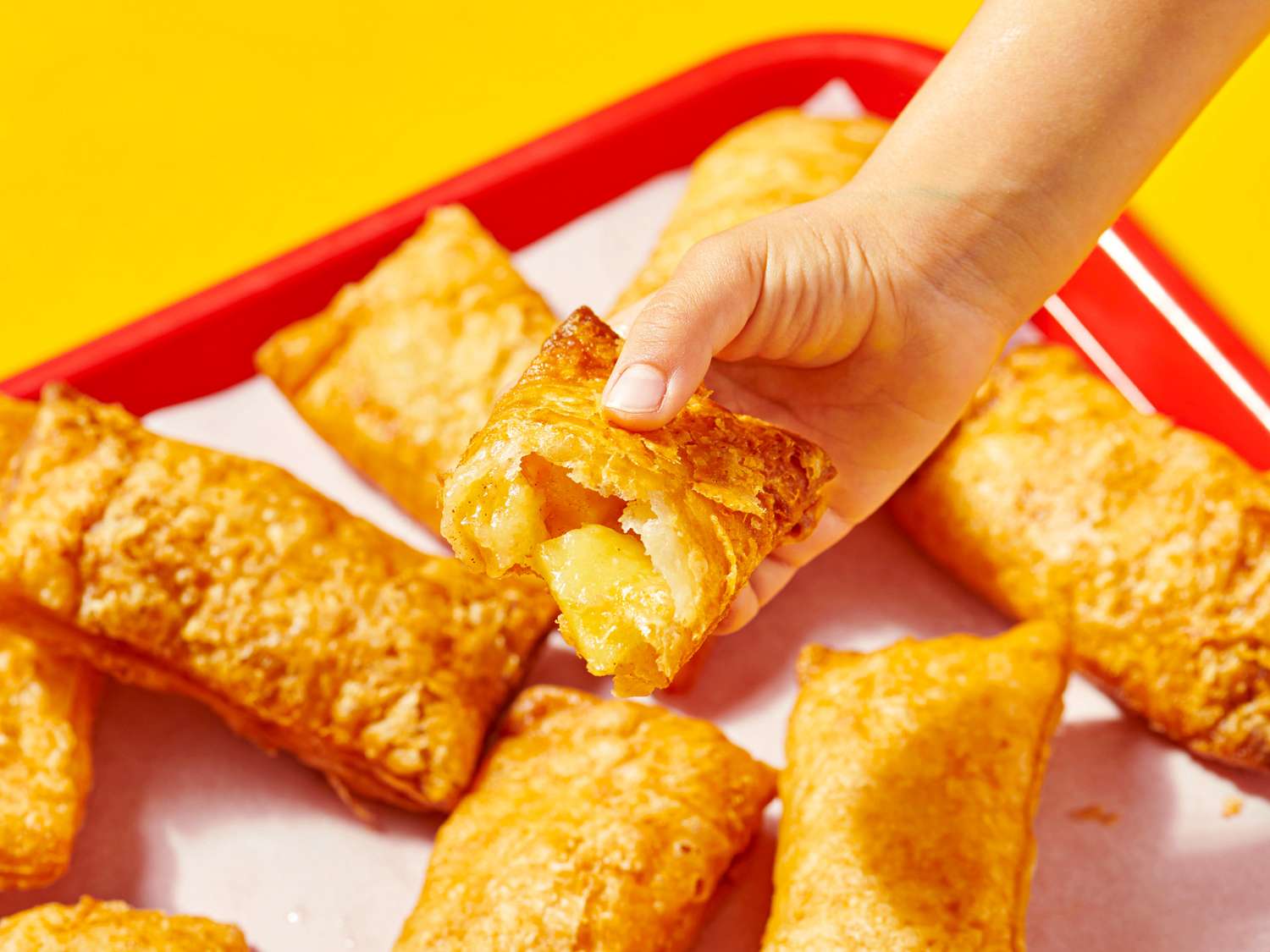 A hand holding a partially broken apple pie above other pies on a tray