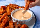 Hand dipping a sweet potato fry into a bowl of sauce sweet potato fries arranged on a plate