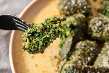 A fork holding a piece of cooked spinach dumpling with more dumplings on a plate in the background
