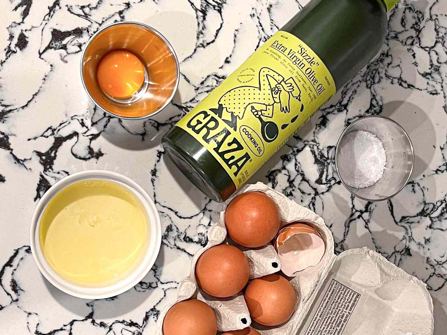 A separated egg, some olive oil, and some salt rest on a marbled countertop. 