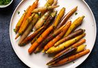 A round white porcelain plate holding multi-colored sous vide glazed carrots. The plate is on a dark blue textured surface and there is a small white bowl holding chopped parsley in the top left corner of the image.
