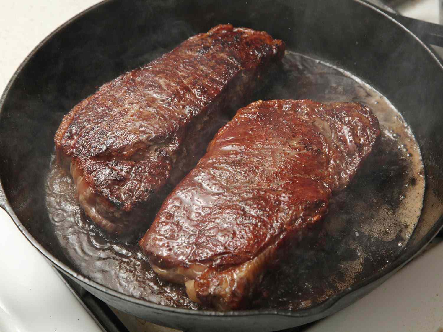 Two thick steaks cooking on a cast-iron skillet with the reverse-sear method
