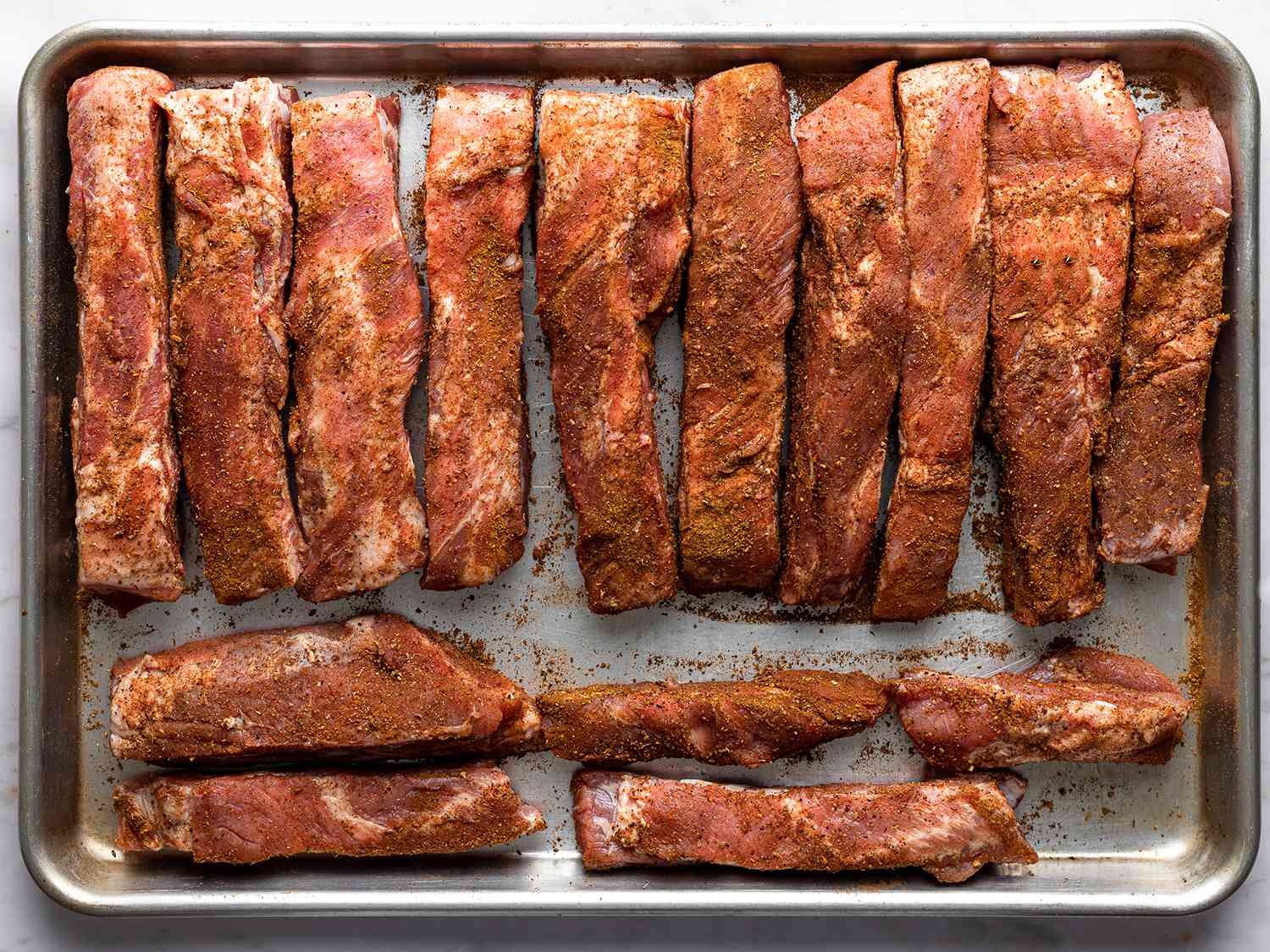 Uncooked ribs covered with spices and laid out on a sheet pan.