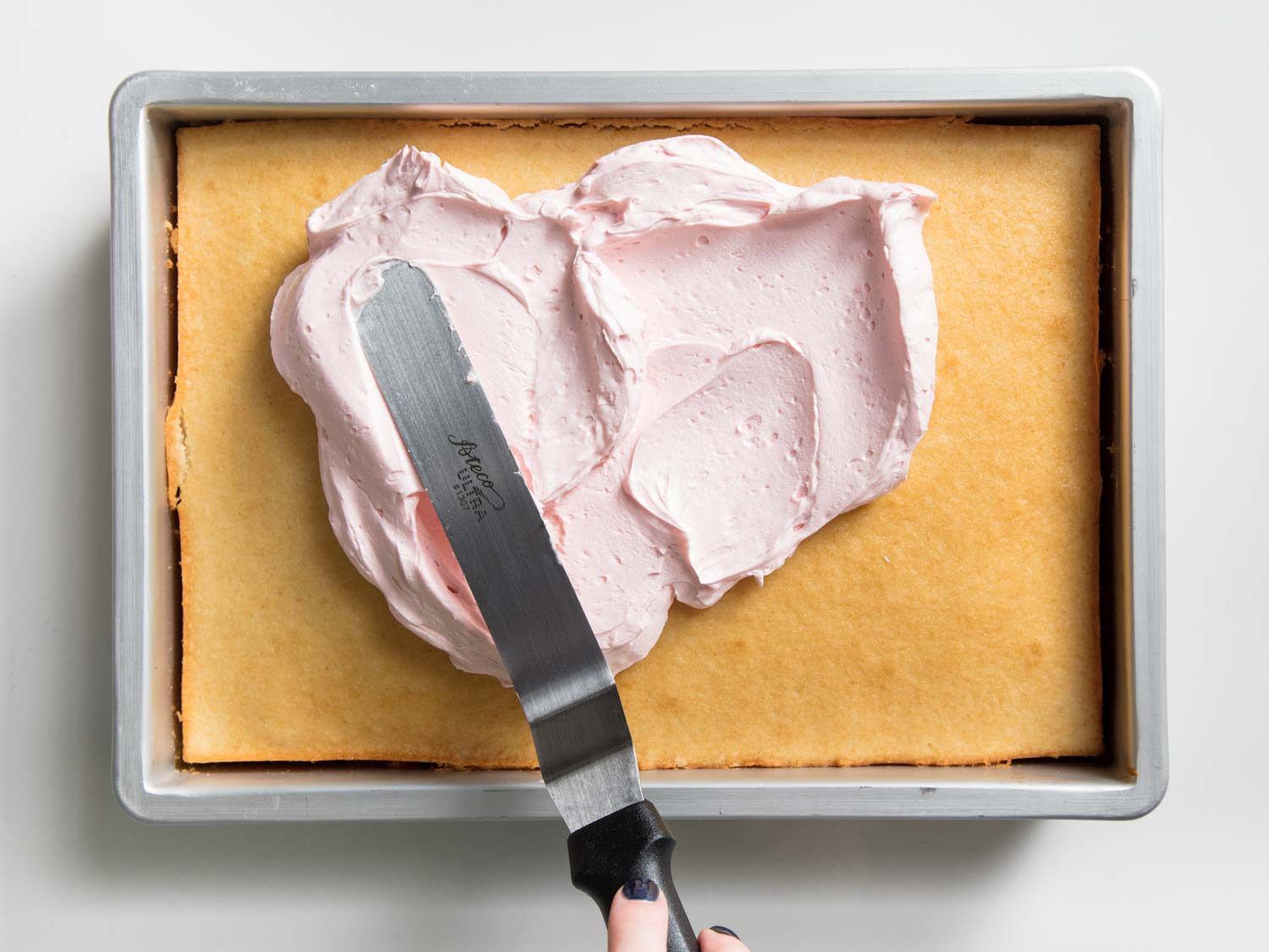 Using an offset spatula to frost a sheet cake with pink American buttercream.