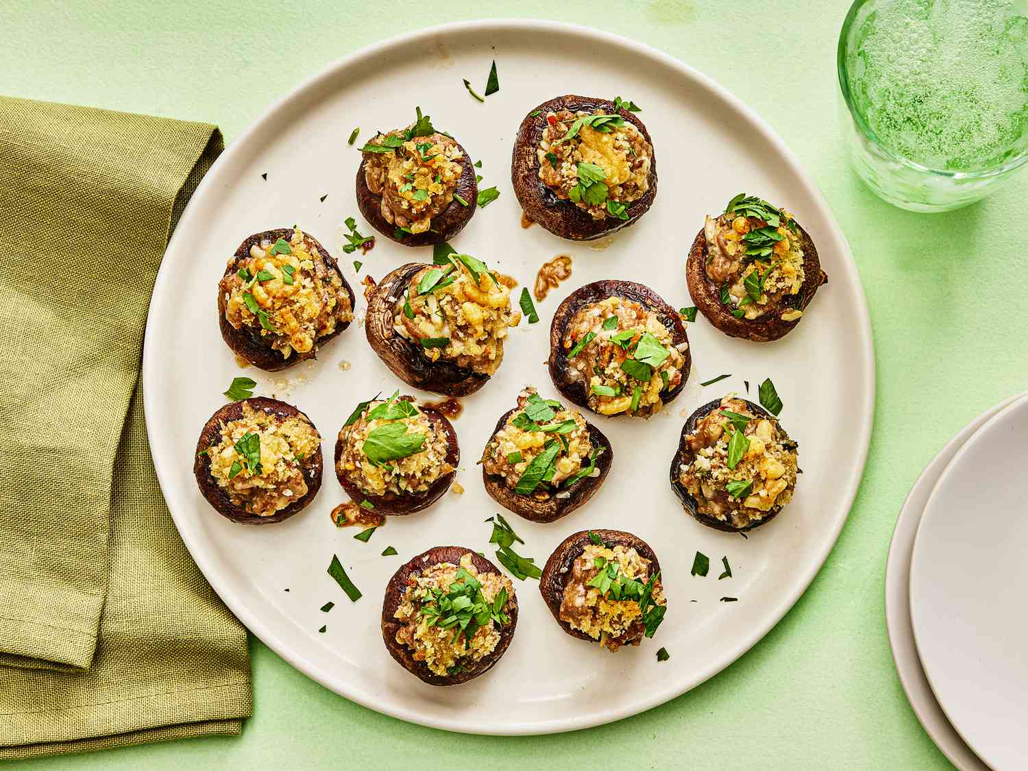 Platter of stuffed mushrooms garnished with parsley on a green surface. Serving plates, a green napkin and a green glass of sparkling water 