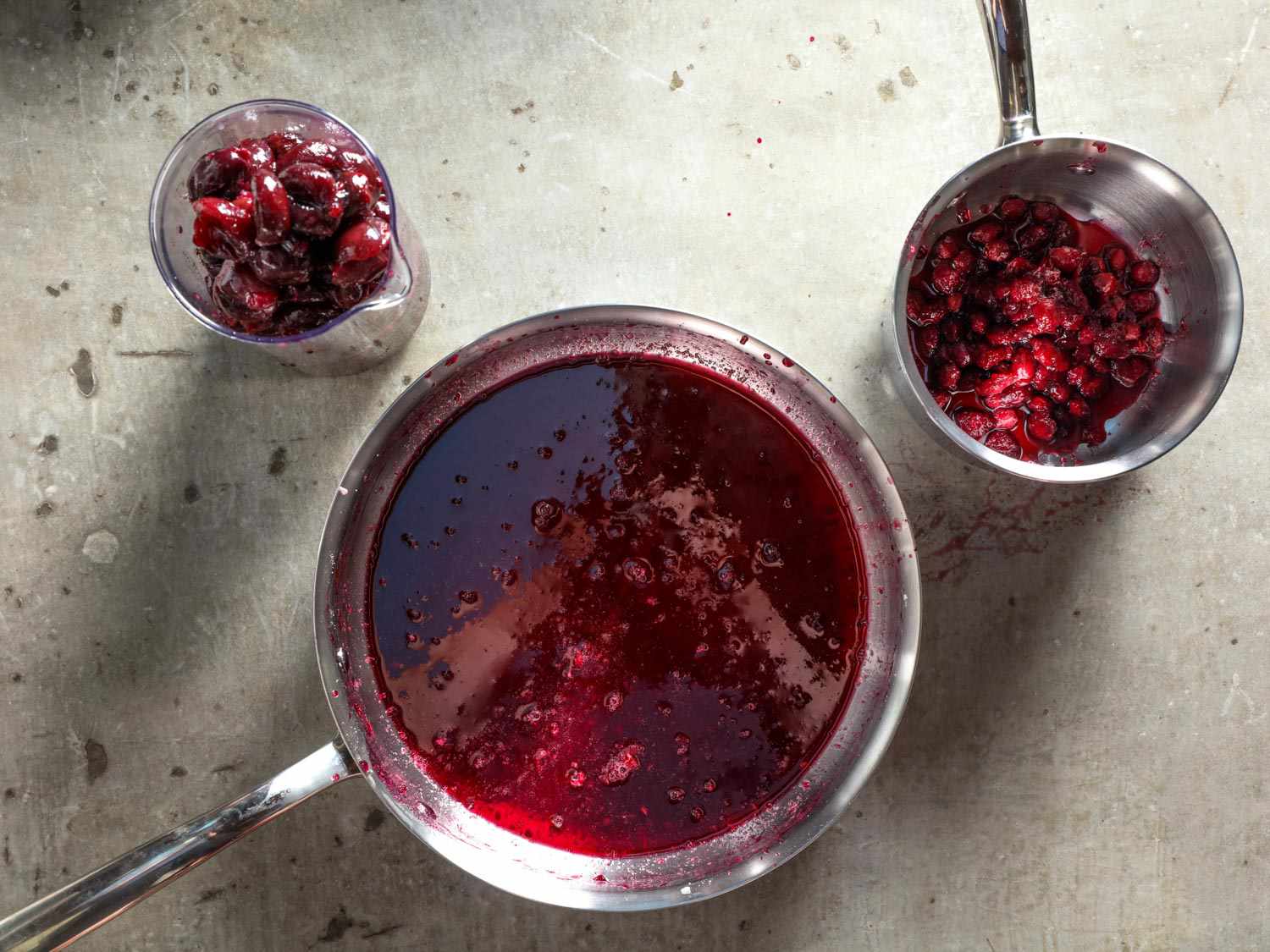 A container of pitted cherries, a saucepan of cherry juice, and a small pan of cherry pits. 