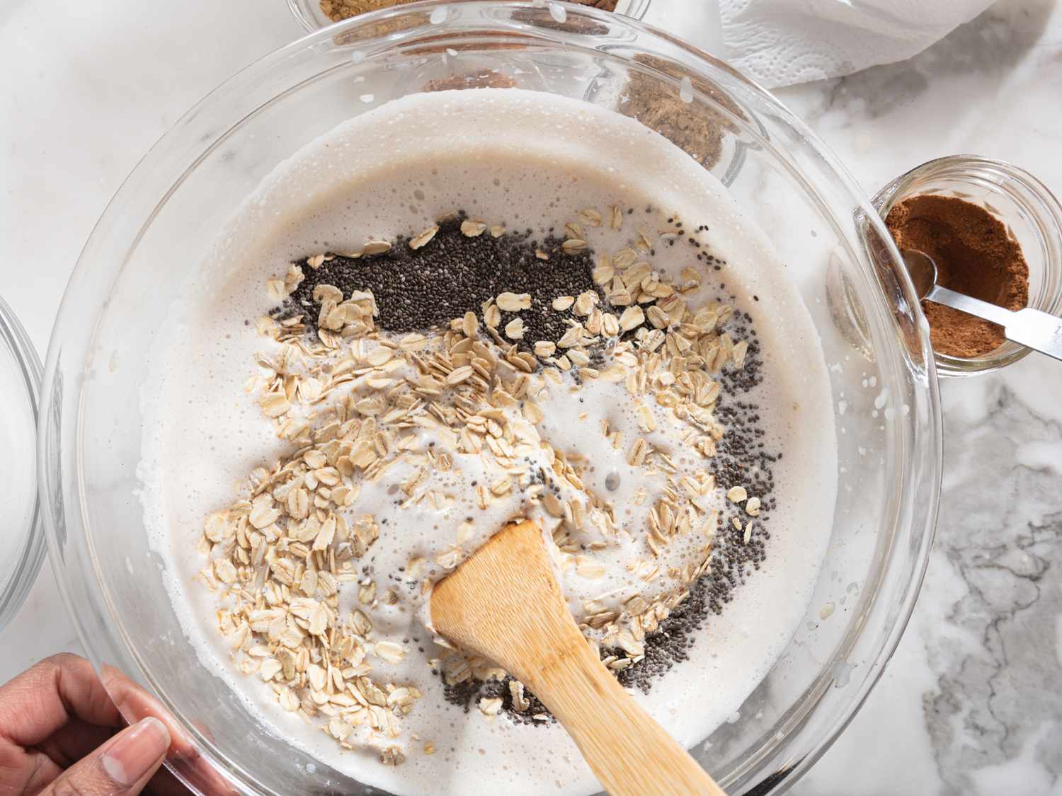 A bowl with oats chia seeds and milk being stirred with a wooden spoon with jars of ingredients visible nearby