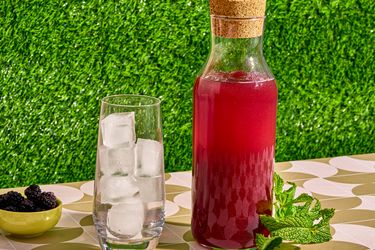 A carafe filled with blackberry liqueur next to a glass of ice, a small bowl of blackberries, and fresh mint sprigs, all on a patterned tablecloth
