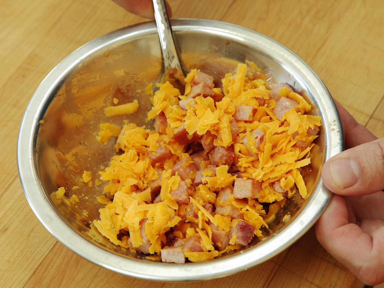 Cubes of browned ham being mixed with shredded cheese in a metal bowl.