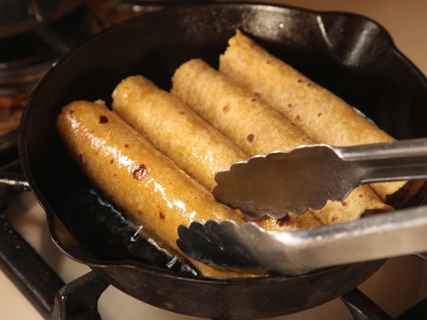 A pair of tongs touching an enchilada frying in a cast iron skillet. 