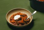 Bowl of beef stew with vegetables and sour cream served with a spoon pot of stew in the background