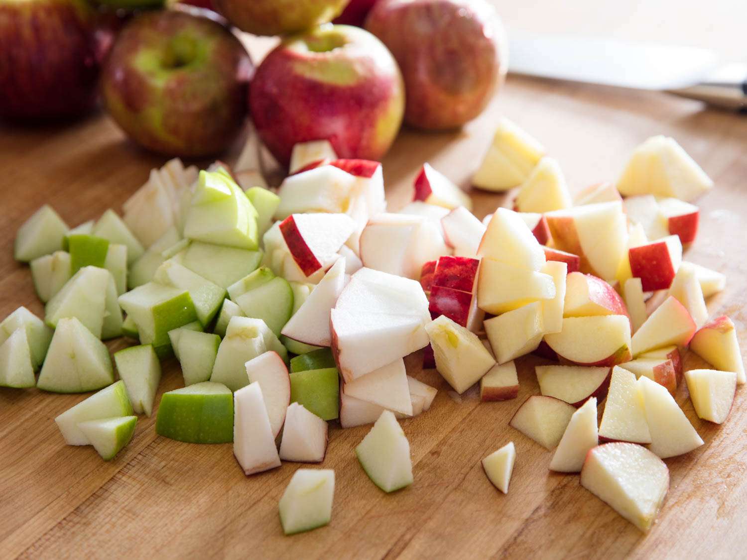 A pile of chopped apple of various varieties and colors, on a wooden surface, with several whole apples in the background.