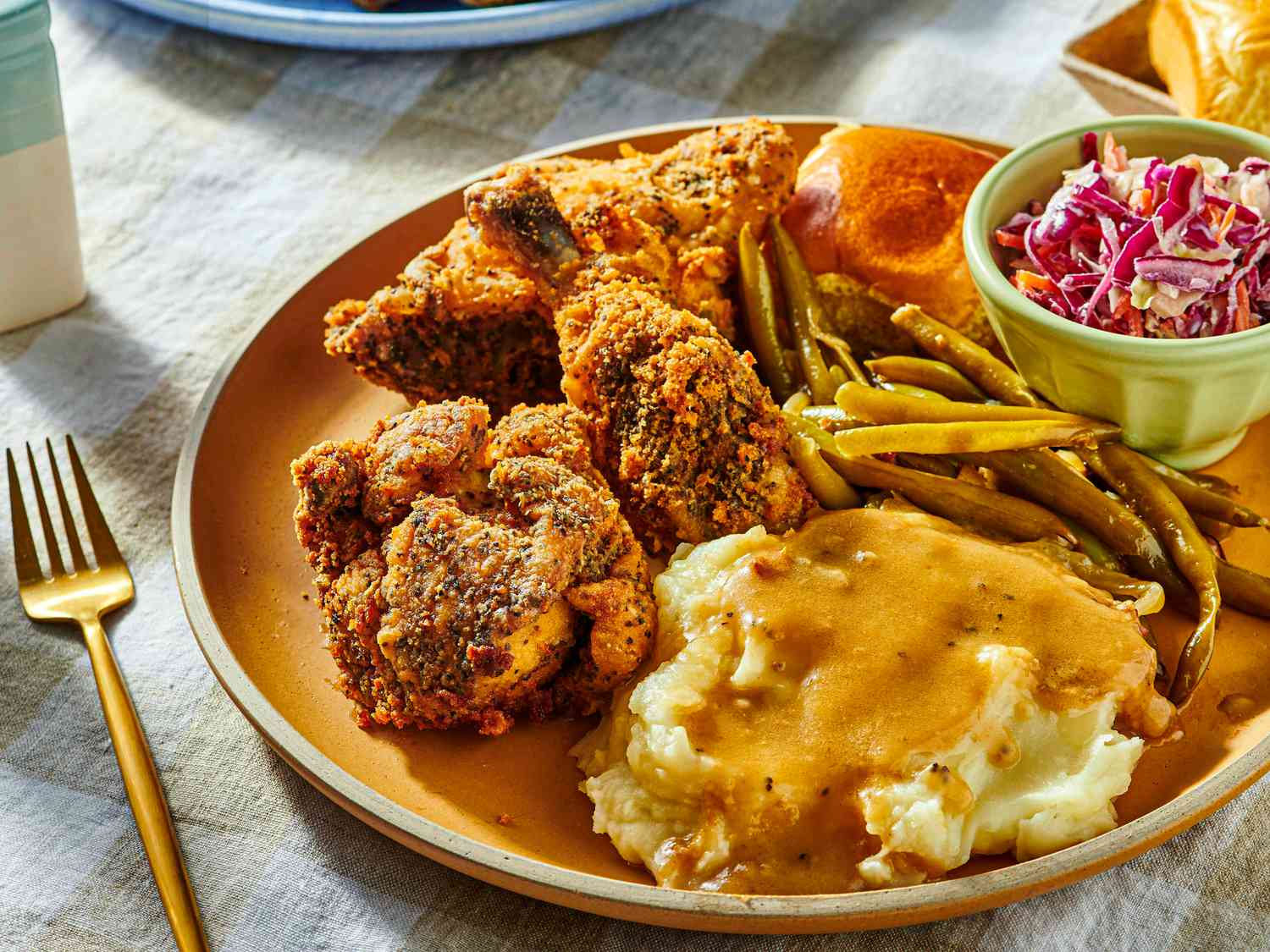 Dish of fried chicken with coleslaw, greenbeans, and mashed potatoes, with a fork and serving dishes in the background